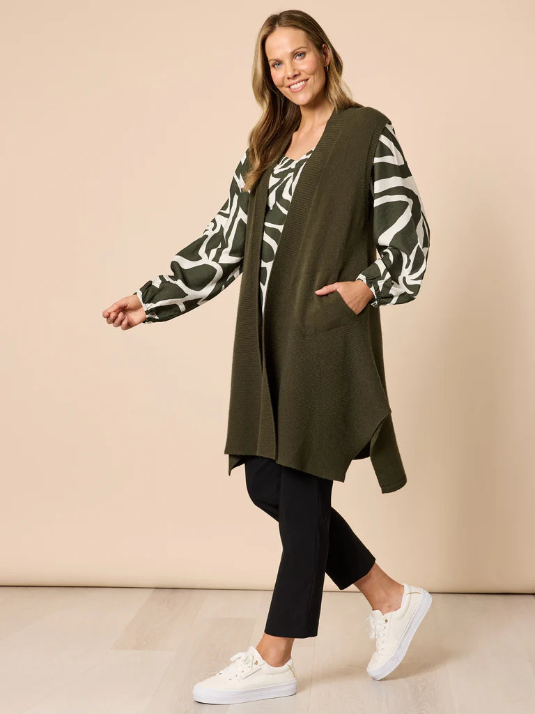 Woman wearing a Clarity green cardigan over a patterned shirt with black pants and white sneakers on a beige background