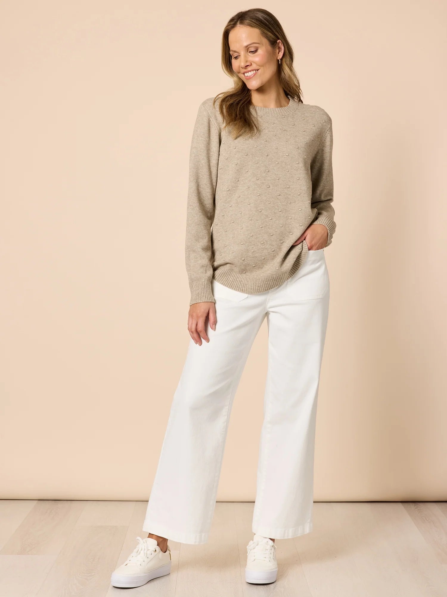 Image of Woman wearing a Clarity beige sweater and white pants against a beige background
