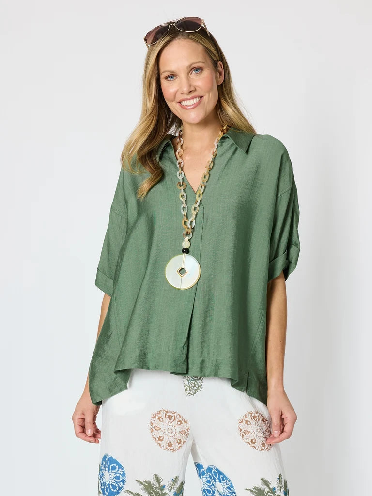 A woman modeling a Clarity sage green short-sleeve button-up shirt with a relaxed, oversized fit, paired with white patterned pants and a chunky gold pendant necklace, against a light gray studio background.
