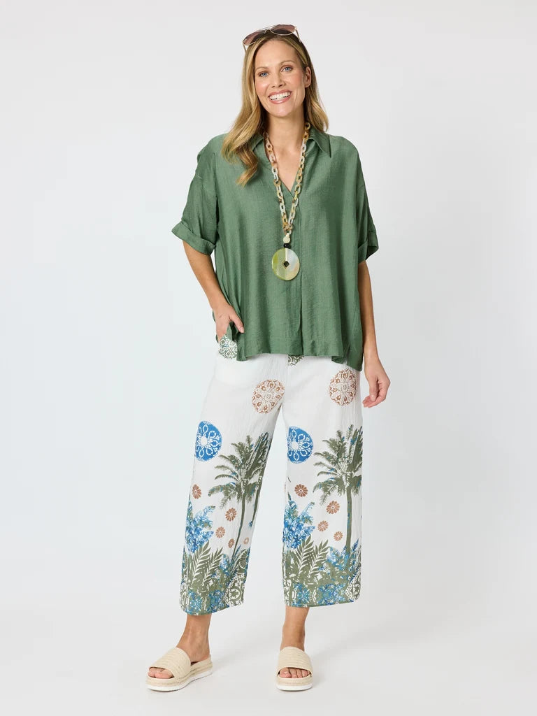 A woman modeling a Clarity green short-sleeve button-up shirt and white wide-leg cropped pants with a blue and green tropical palm tree print, styled with a long gold pendant necklace and beige slide sandals against a light gray studio background.