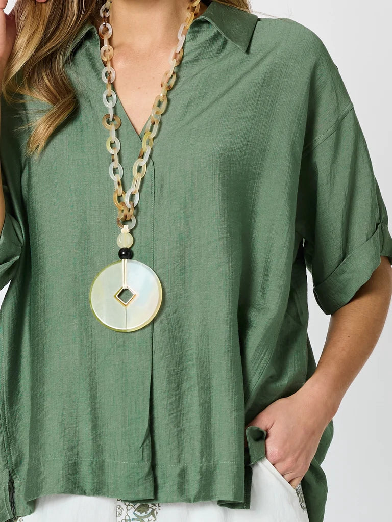 A woman wearing a Clarity sage green linen button-down shirt and a chunky multi-strand necklace with a large round shell pendant.