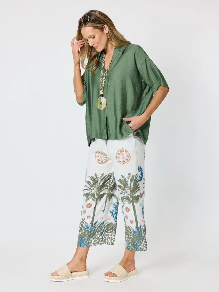 Image of A woman modeling a Clarity green short-sleeve blouse and white cropped pants with a blue and green palm tree print, styled with beige slide sandals and a long pendant necklace against a light gray studio background.