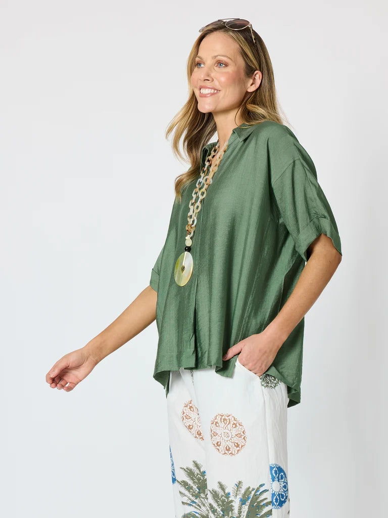 Image of A woman modeling a Clarity loose, short-sleeve sage green top paired with white patterned pants, standing against a light gray studio background.