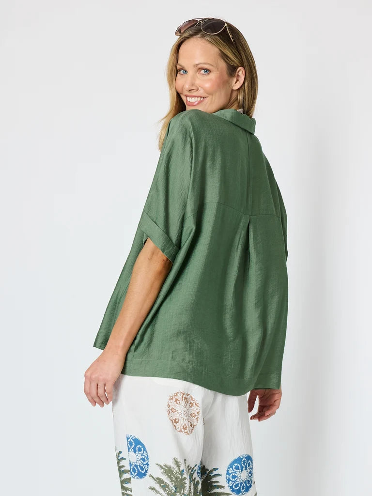 Image of A woman wearing a Clarity olive green short-sleeve button-up shirt and white tropical-print pants, photographed against a plain light gray studio background.