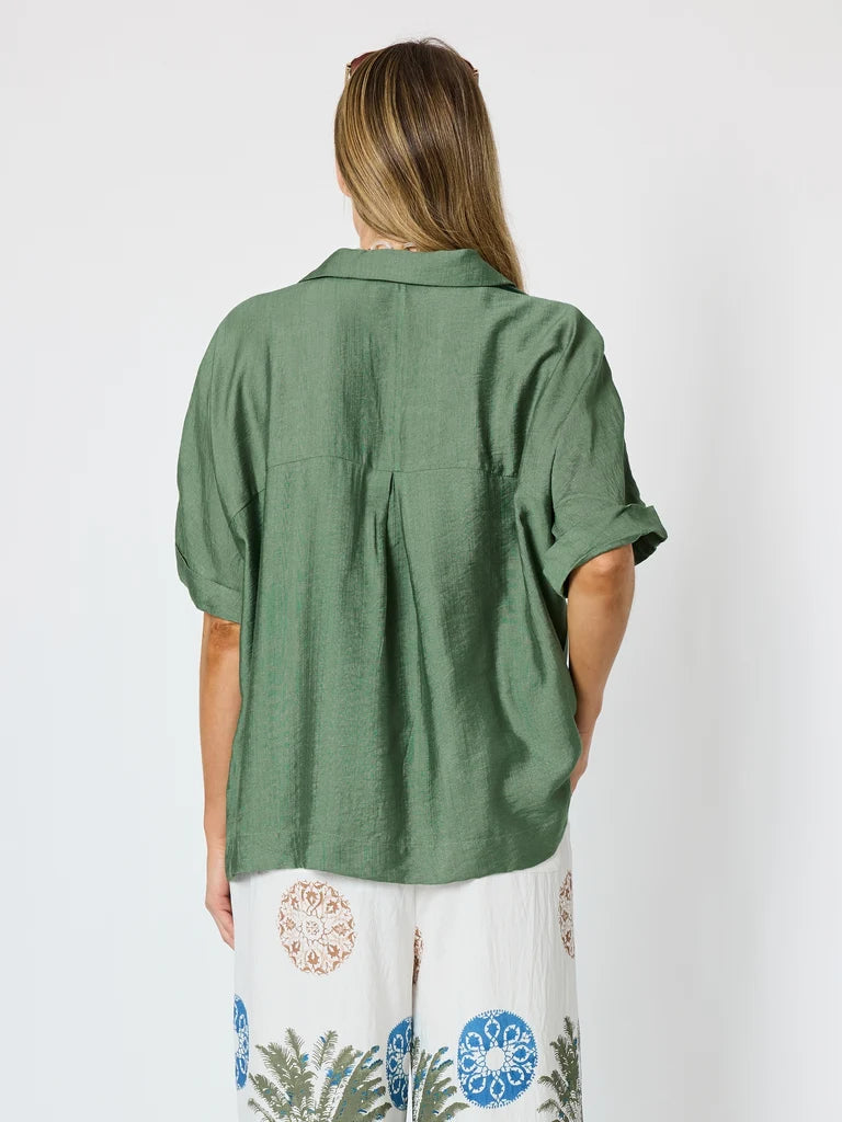 Image of A woman shown from the back wearing a Clarity sage green short-sleeve linen shirt and white pants with blue and orange botanical patterns, against a plain light gray background.