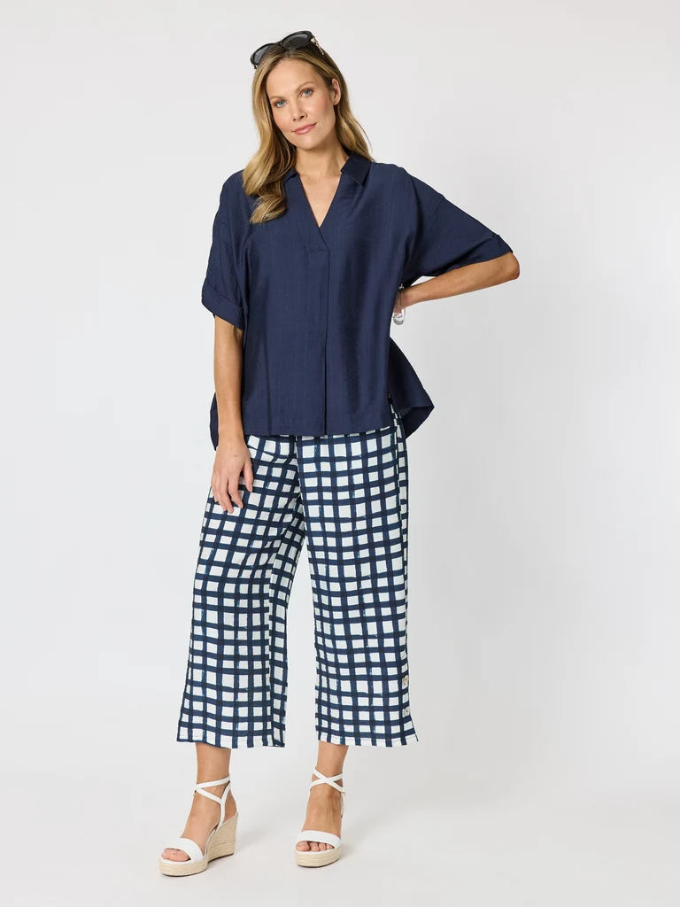 Image of A woman modeling a Clarity navy blue short-sleeve button-up shirt and navy and white gingham cropped pants, paired with white wedge sandals, against a light gray studio background.