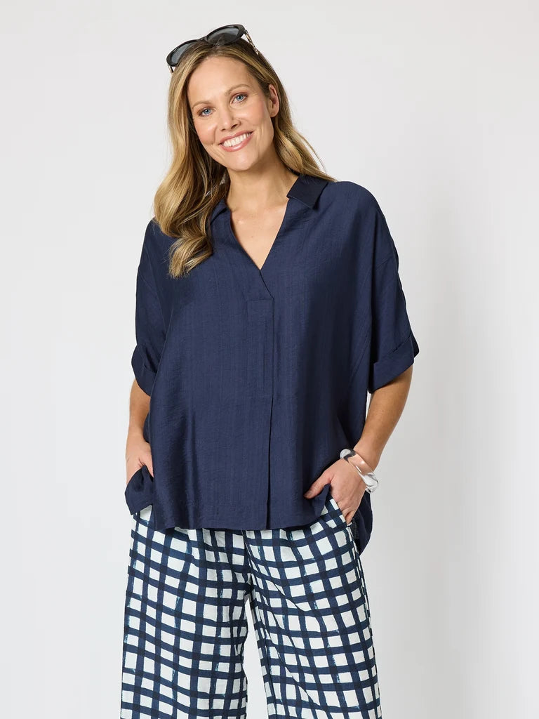 Image of A woman modeling a Clarity navy blue short-sleeve button-up shirt with a relaxed fit, paired with navy and white checkered pants, against a plain light gray background.