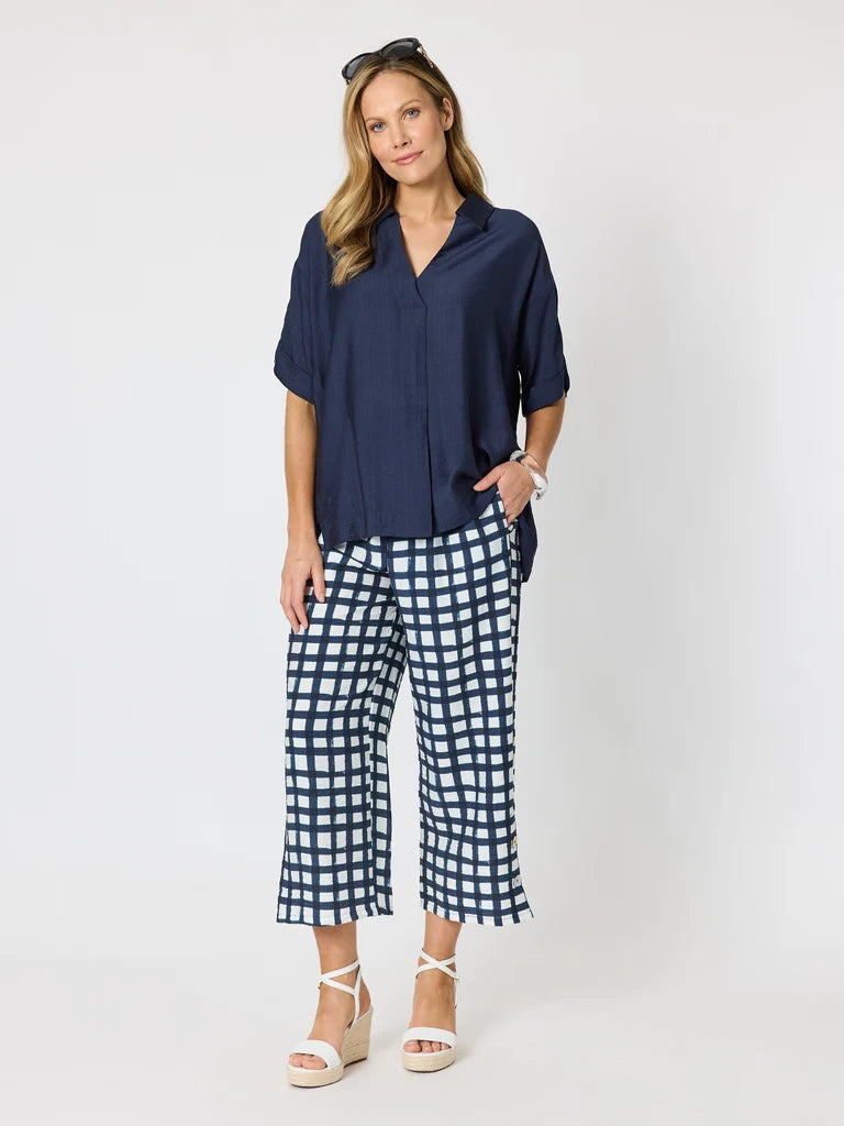 Image of A woman modeling a Clarity navy blue short-sleeve button-up shirt paired with navy and white gingham cropped pants and white wedge sandals, photographed against a light gray studio background.