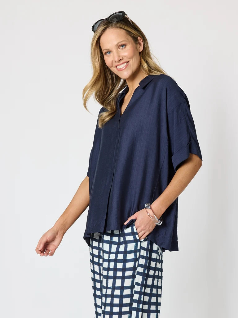Image of A woman modeling a Clarity navy blue short-sleeve button-up shirt with a relaxed fit, paired with navy and white checkered pants, against a plain light gray background.
