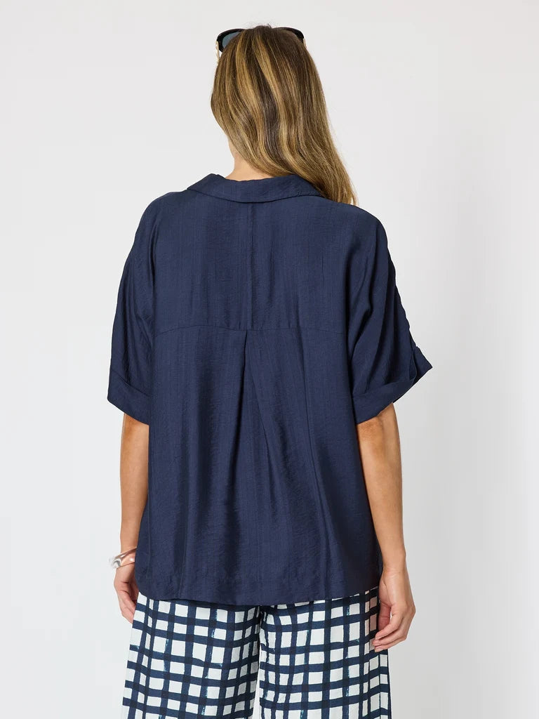 Image of A woman viewed from the back wearing a Clarity navy blue short-sleeve button-up shirt and navy-and-white checkered pants, against a plain light gray studio background.