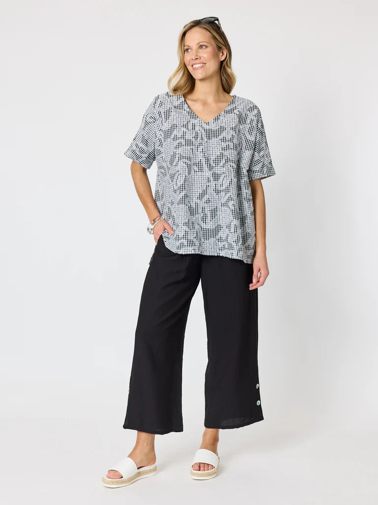 Image of A woman modeling a Clarity short-sleeve V-neck top with a gray and white abstract pattern, paired with black wide-leg cropped pants and white slide sandals, against a light gray studio background.