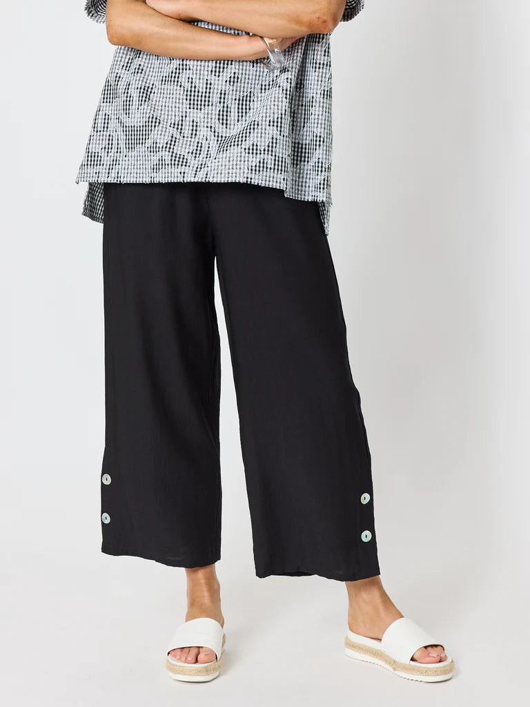 A woman wearing Clarity black wide-leg cropped pants with decorative buttons on the lower hems, paired with a black-and-white patterned top and white slide sandals, against a light gray studio background.