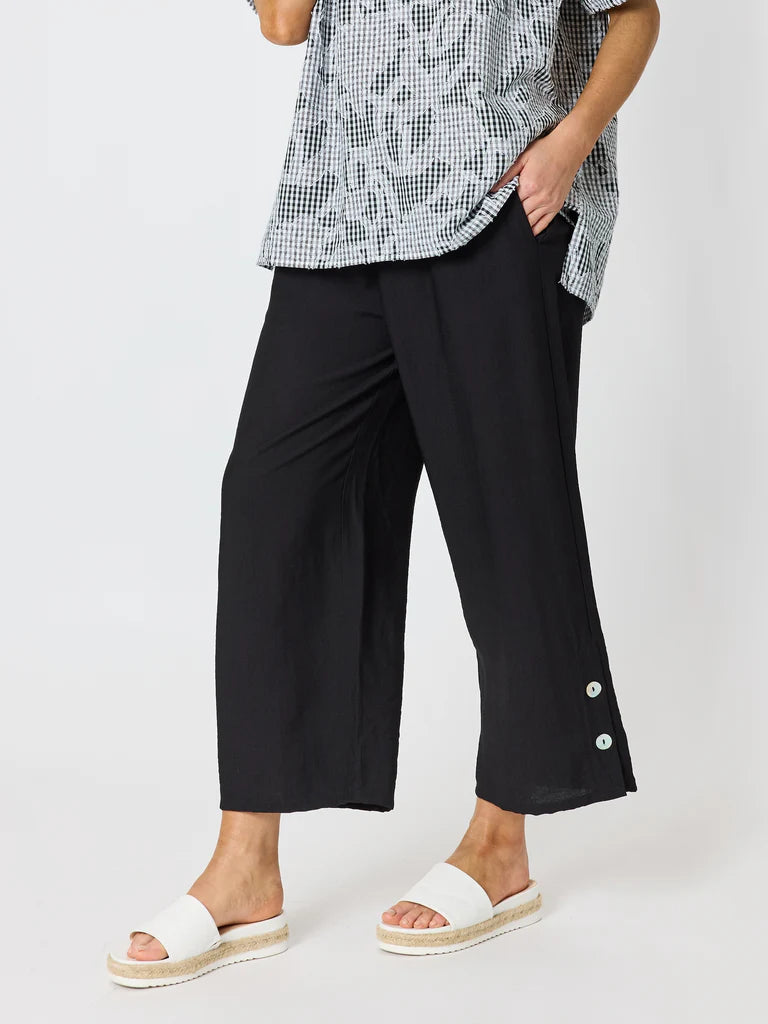 A woman wearing Clarity black wide-leg cropped pants with decorative buttons on the lower hems, paired with a black-and-white patterned top and white slide sandals, against a light gray studio background.
