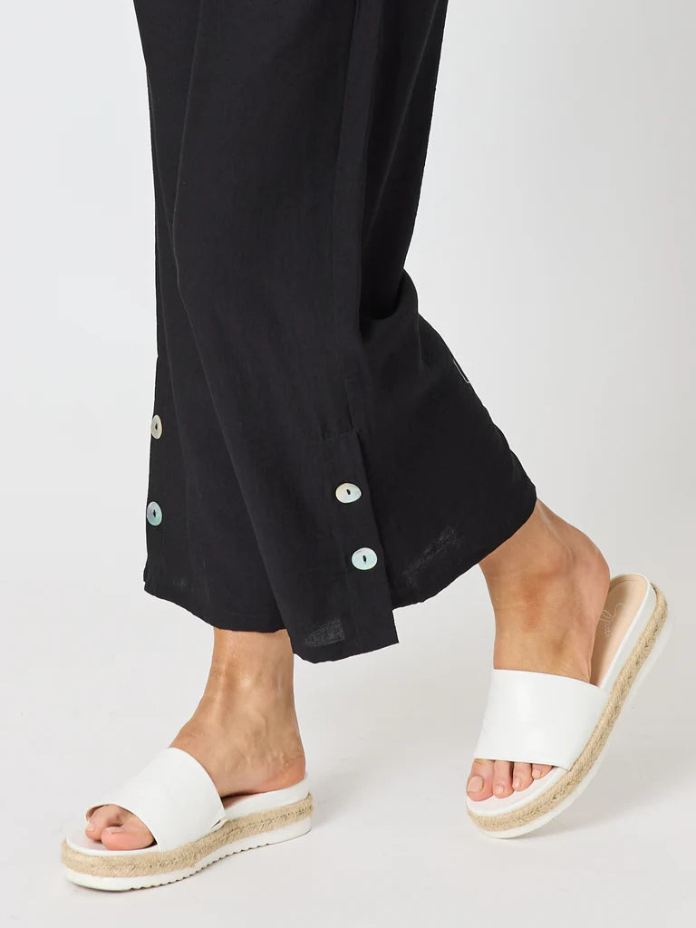 Image of A close-up of a person's feet wearing Clarity black wide-leg cropped trousers with decorative buttons and white espadrille-style slide sandals with a braided jute platform sole, against a light gray studio background.