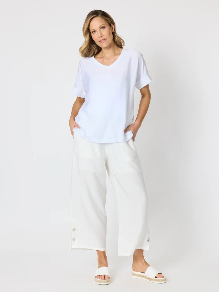 Image of A woman modeling a white short-sleeve V-neck top and Clarity white wide-leg cropped pants with button details at the hem, paired with white slide sandals, against a light gray studio background.