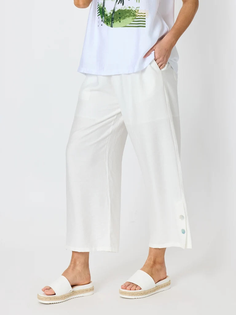 Image of A model wearing Clarity white wide-leg cropped pants with side buttons, styled with a white graphic t-shirt and white slide sandals on a light gray studio background.