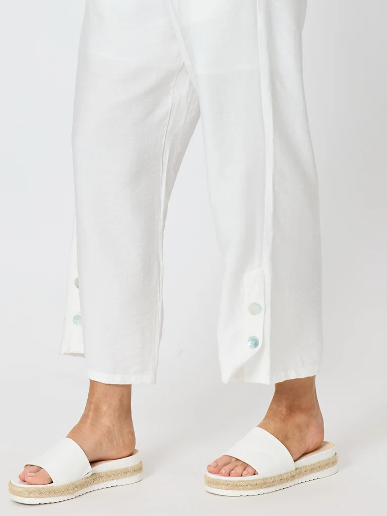 Clarity white cropped pants with decorative buttons on the lower hems, shown on a model wearing white slide sandals with espadrille-style soles against a light gray studio background.