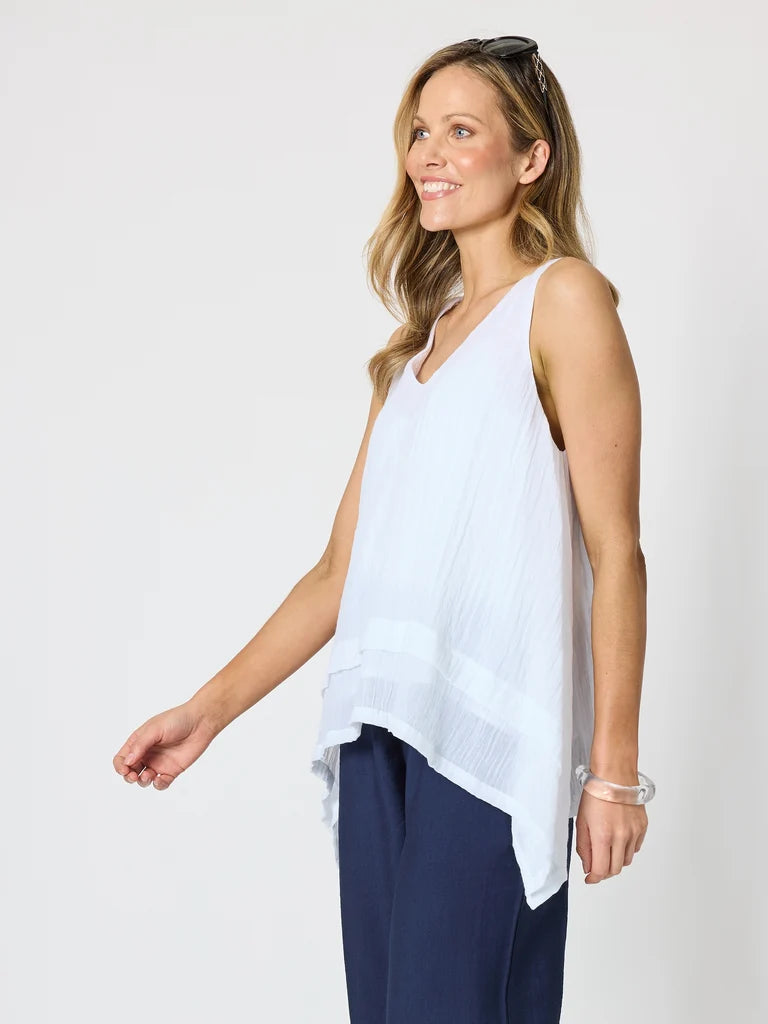 Image of A woman wearing a Clarity white sleeveless V-neck top with a high-low hem and navy blue pants, standing against a light gray studio background.