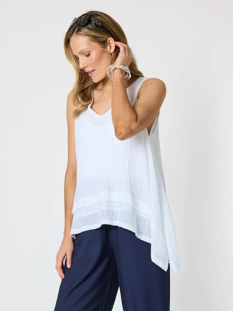 Image of A woman wearing a Clarity white sleeveless V-neck top with a tiered hem and navy blue pants, photographed against a light gray studio background.