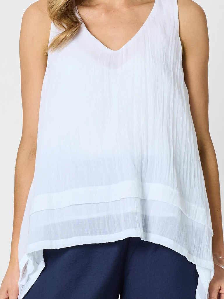 A woman wearing a Clarity white sleeveless V-neck top with a textured, crinkled fabric and a tiered hem, paired with navy blue pants.