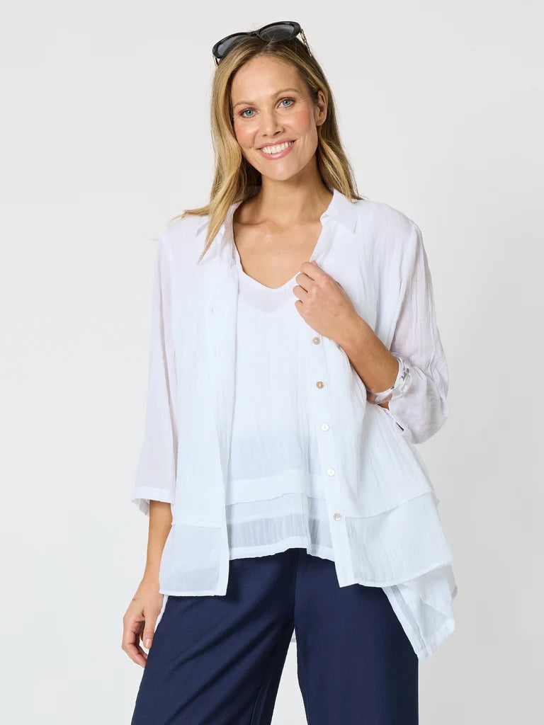 Image of A woman modeling a Clarity white tiered button-front blouse with three-quarter sleeves, layered over a white V-neck top and paired with navy pants, against a light gray studio background.