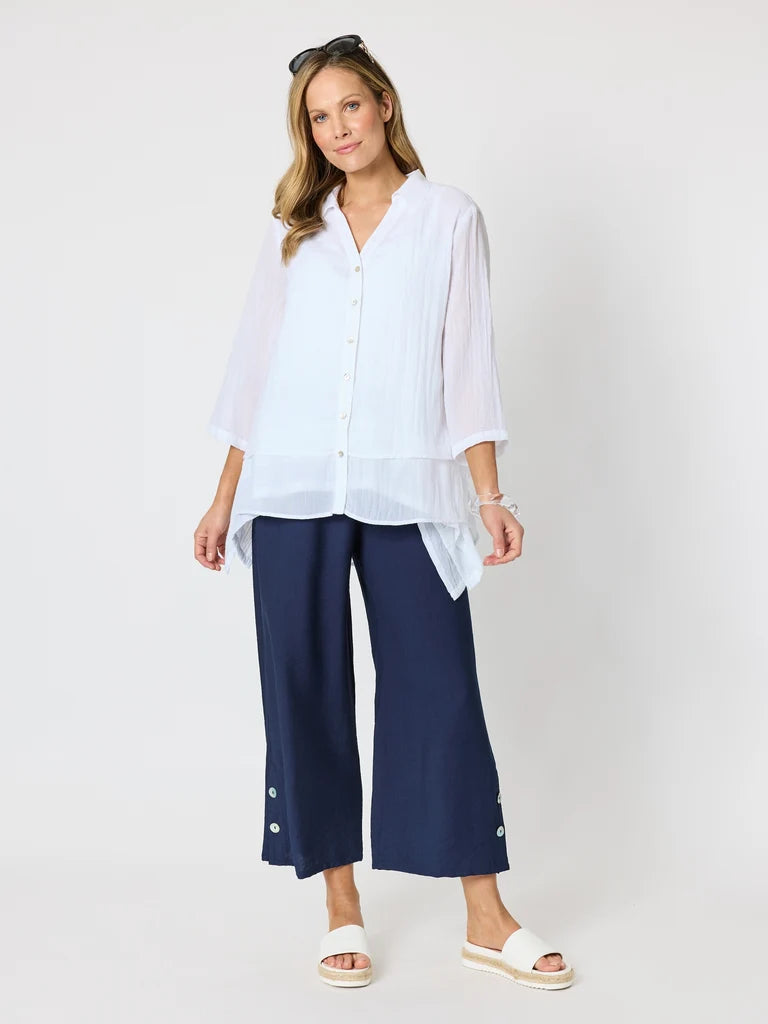 Image of A woman modeling a Clarity white sheer long-sleeve button-up shirt paired with navy blue cropped wide-leg pants and white slide sandals, photographed against a light gray studio background.