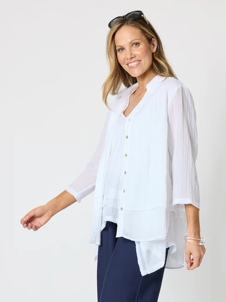 Image of A woman modeling a Clarity white long-sleeve button-up blouse with a V-neck and layered ruffle hem, paired with navy pants against a light gray studio background.