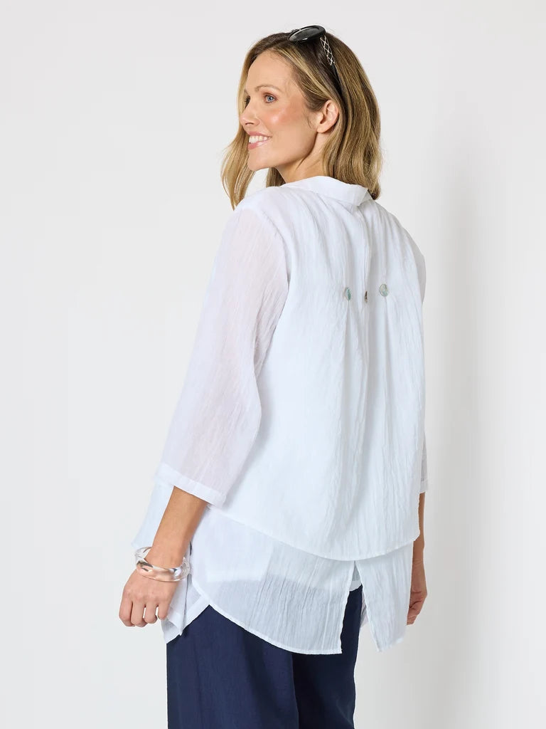 A woman wearing a Clarity white, semi-sheer three-quarter sleeve blouse with a high-low hem and button details on the back, paired with navy pants, photographed against a plain white studio background.