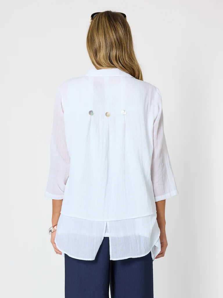 A woman viewed from the back wearing a Clarity white lightweight blouse with three decorative buttons on the back and navy blue pants, against a plain light background.