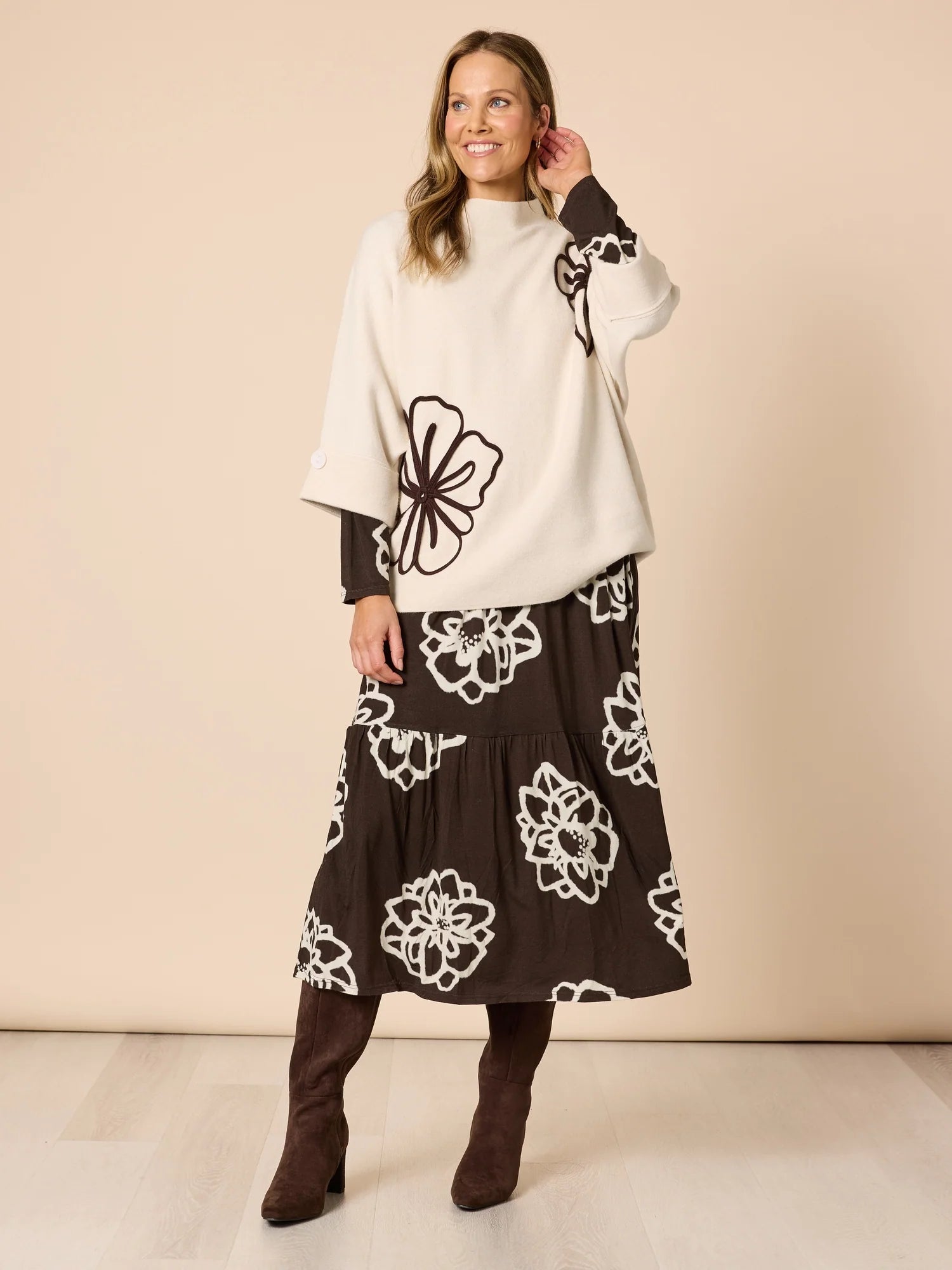 Image of Woman wearing a white sweater with Clarity black floral patterns and a brown skirt with white floral patterns on a beige background.