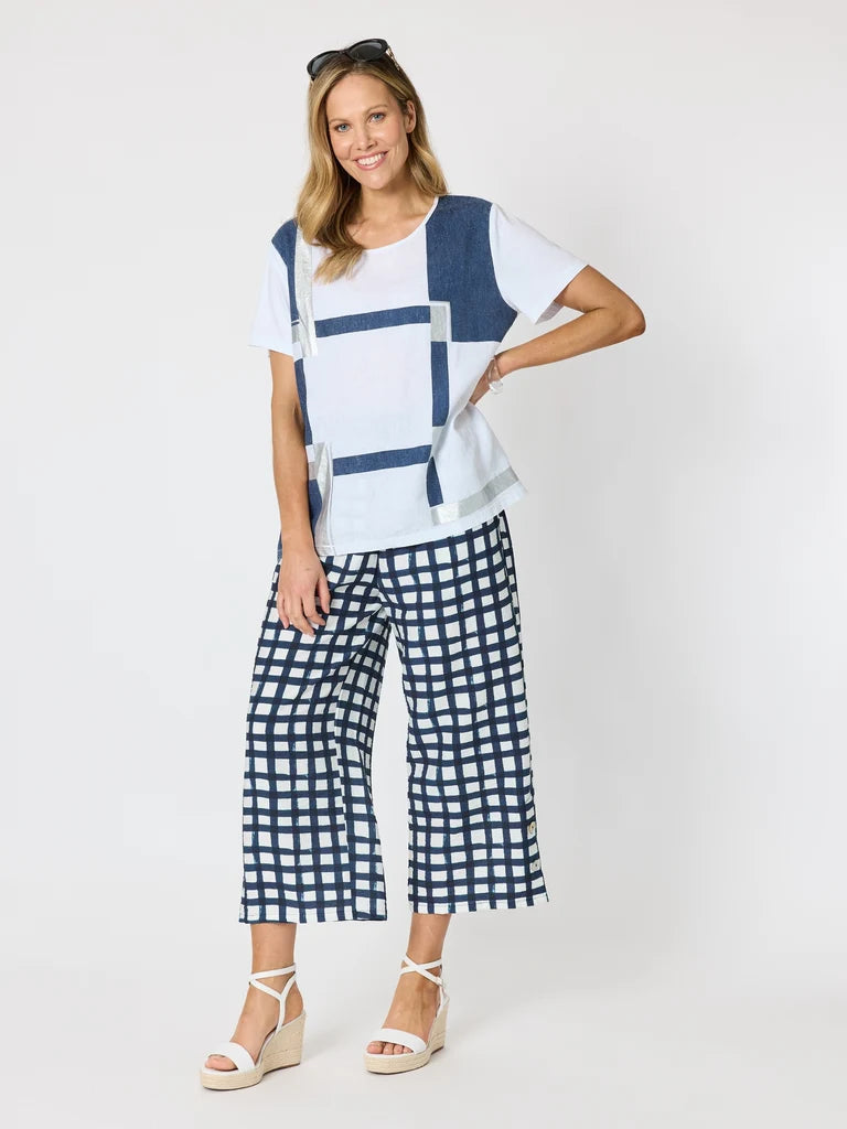 Image of A woman modeling a Clarity white short-sleeve top with blue geometric block patterns and matching blue-and-white checkered cropped pants, paired with white wedge sandals, against a plain light gray studio background.