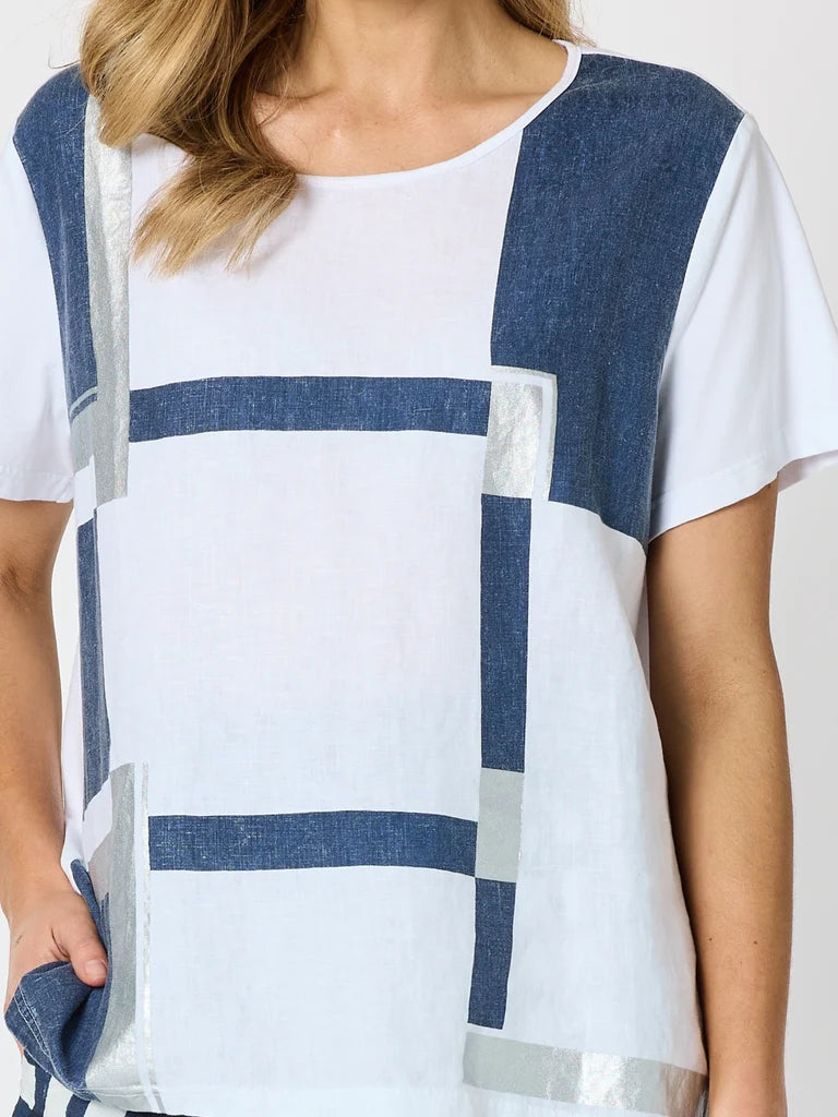 Image of A close-up of a woman wearing a Clarity white short-sleeve top with a blue geometric block pattern and metallic silver stripe accents.