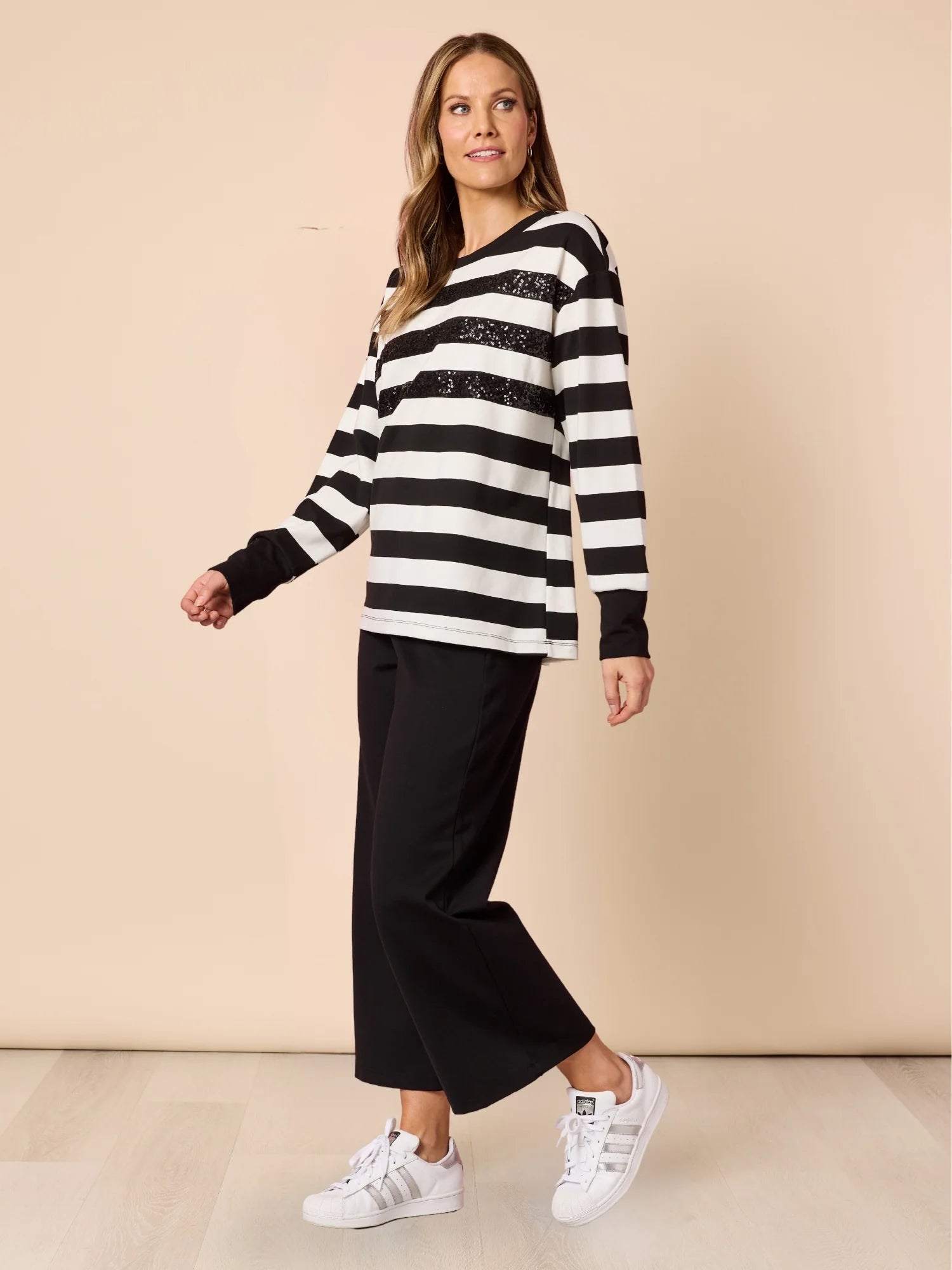 Woman wearing a black and white striped sweater with Clarity black pants and white sneakers on a beige background