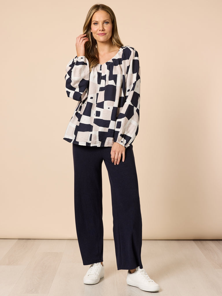 Image of Woman wearing a patterned blouse and Clarity navy pants against a beige background