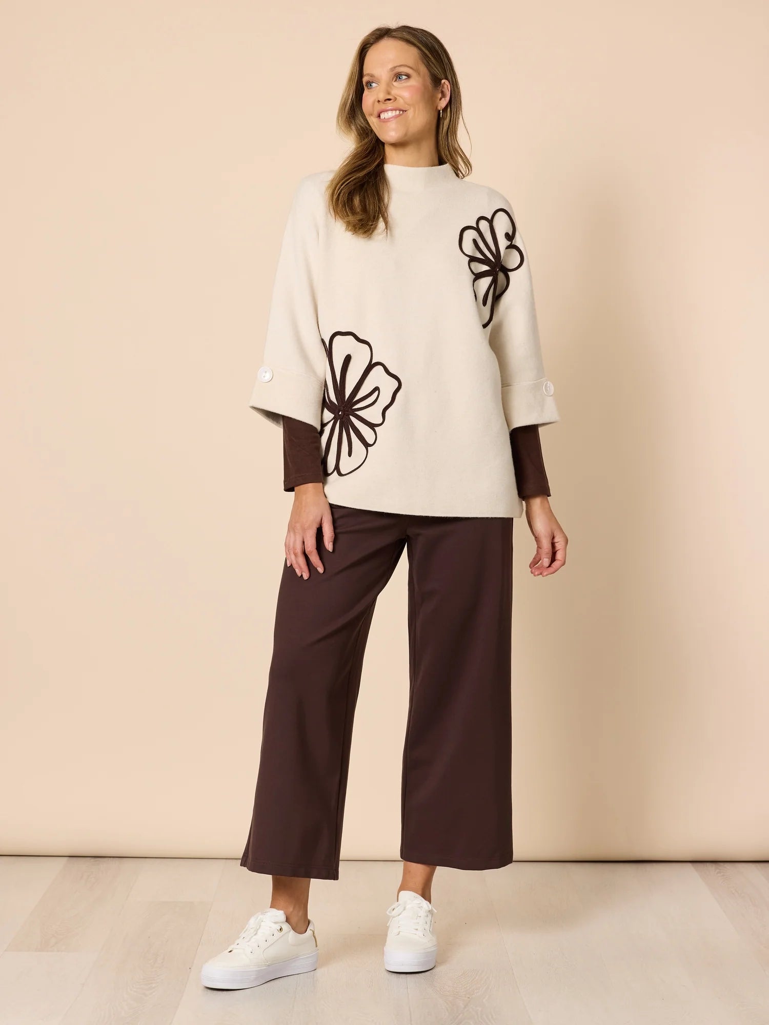 Woman wearing a Clarity cream sweater with black floral designs and brown pants on a beige background