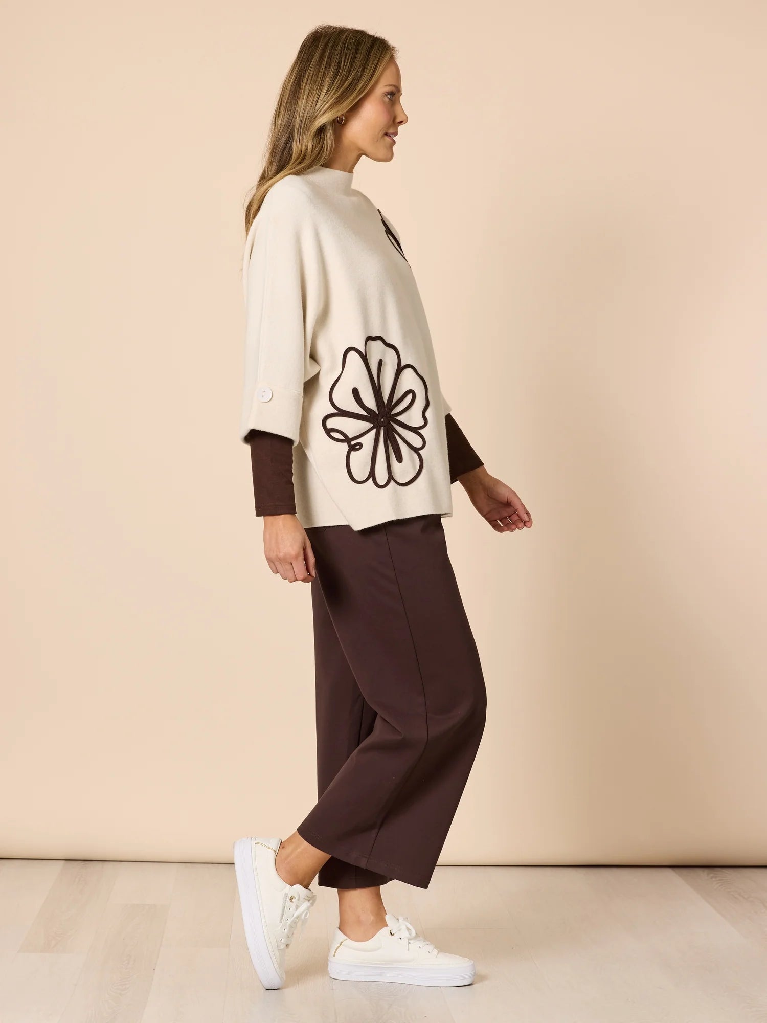 Image of Woman wearing a Clarity beige sweater with a black flower design and brown pants on a beige background