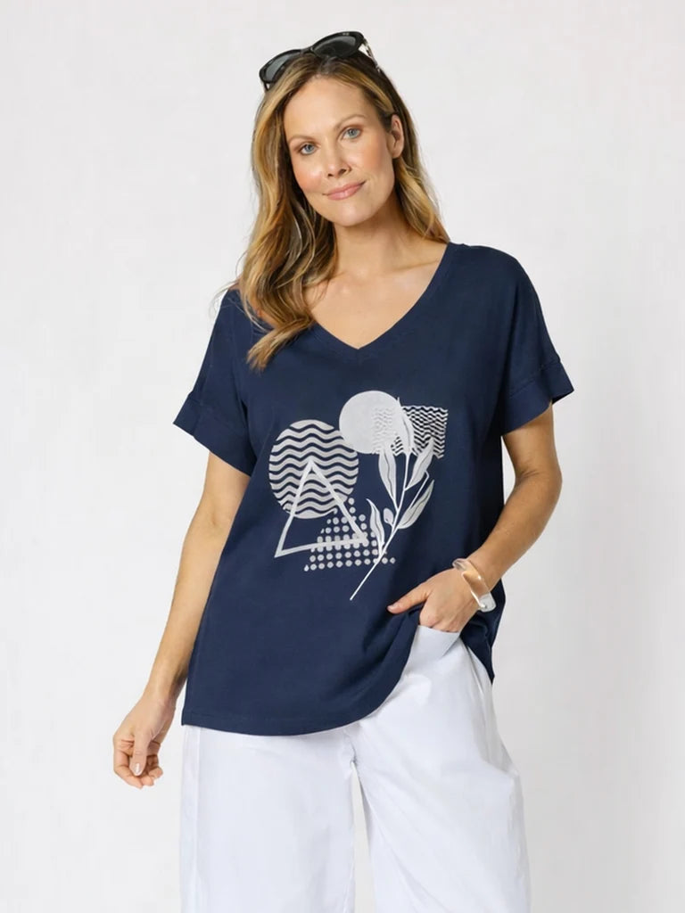 Image of A woman modeling a Clarity navy blue V-neck short-sleeve t-shirt with a white graphic print of a sun, moon, and geometric shapes, paired with white pants against a light gray background.