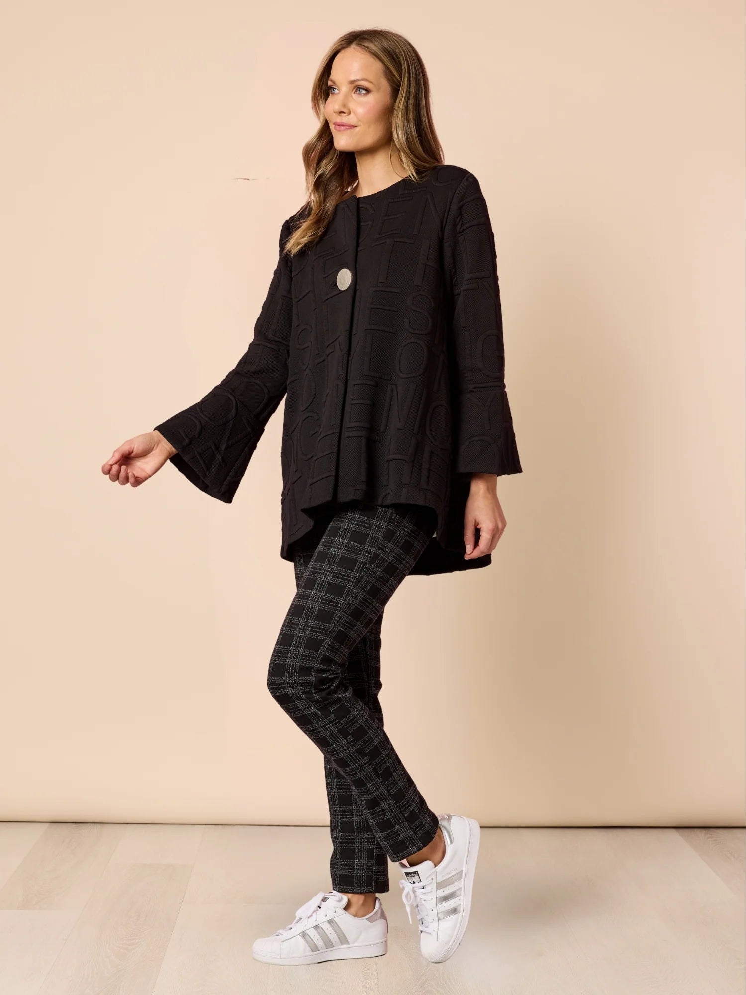 Woman wearing a Clarity black jacket with plaid pants and white sneakers on a beige background