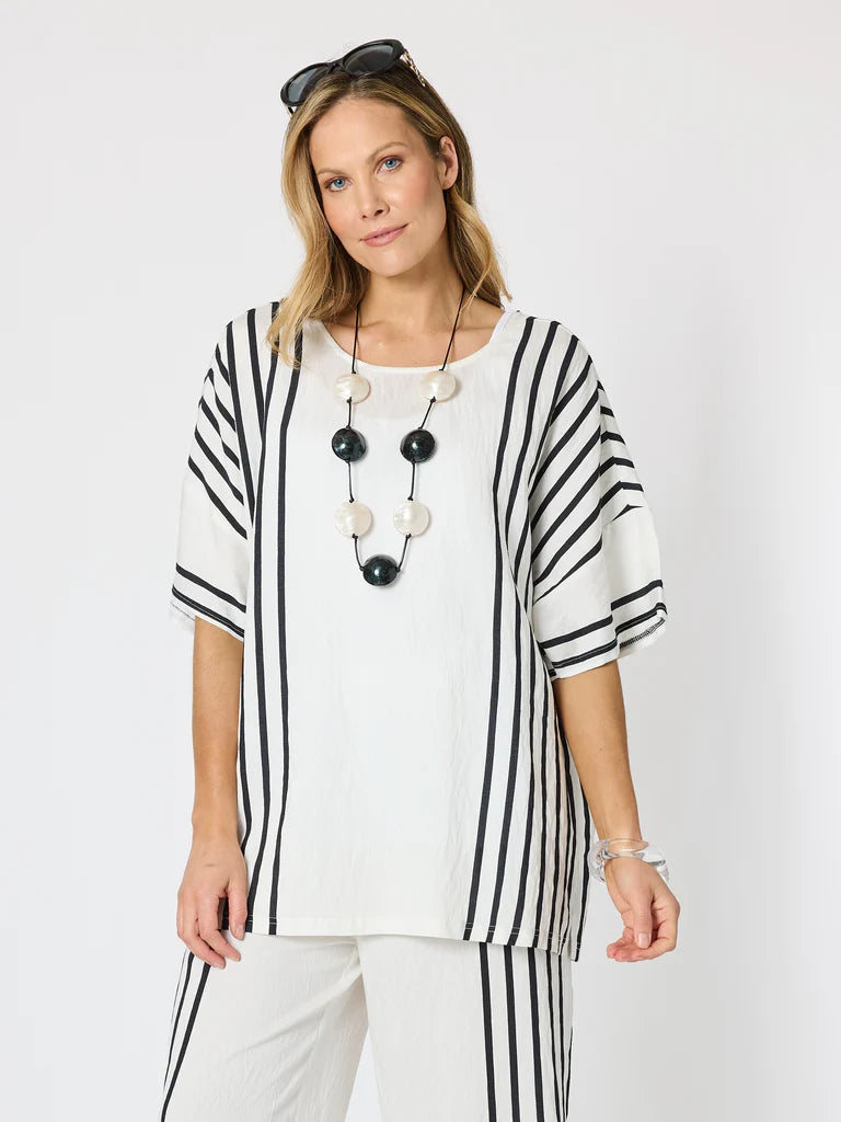 Image of A woman modeling a Clarity white short-sleeve top with black vertical stripes and matching striped pants, accessorized with a long black and white beaded necklace and sunglasses on her head, against a plain light gray background.