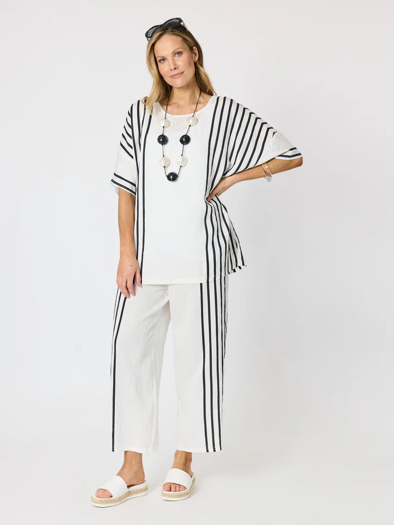 Image of A woman modeling a Clarity white two-piece outfit featuring a short-sleeve top with black vertical stripes and matching wide-leg cropped pants with black side stripes, styled with a long beaded necklace and white slide sandals against a light gray studio background.