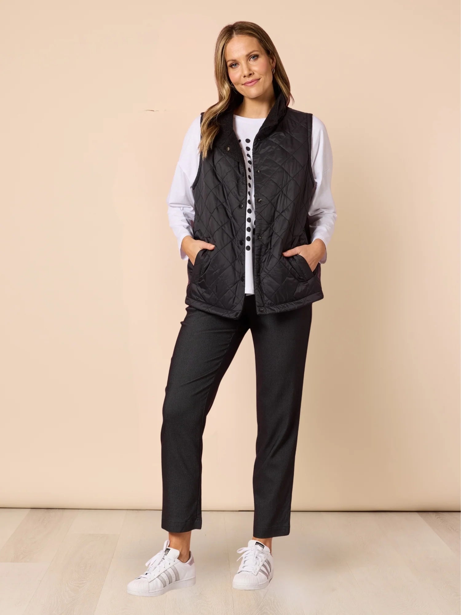 Woman wearing a Clarity black quilted vest over a white shirt with black pants on a beige background