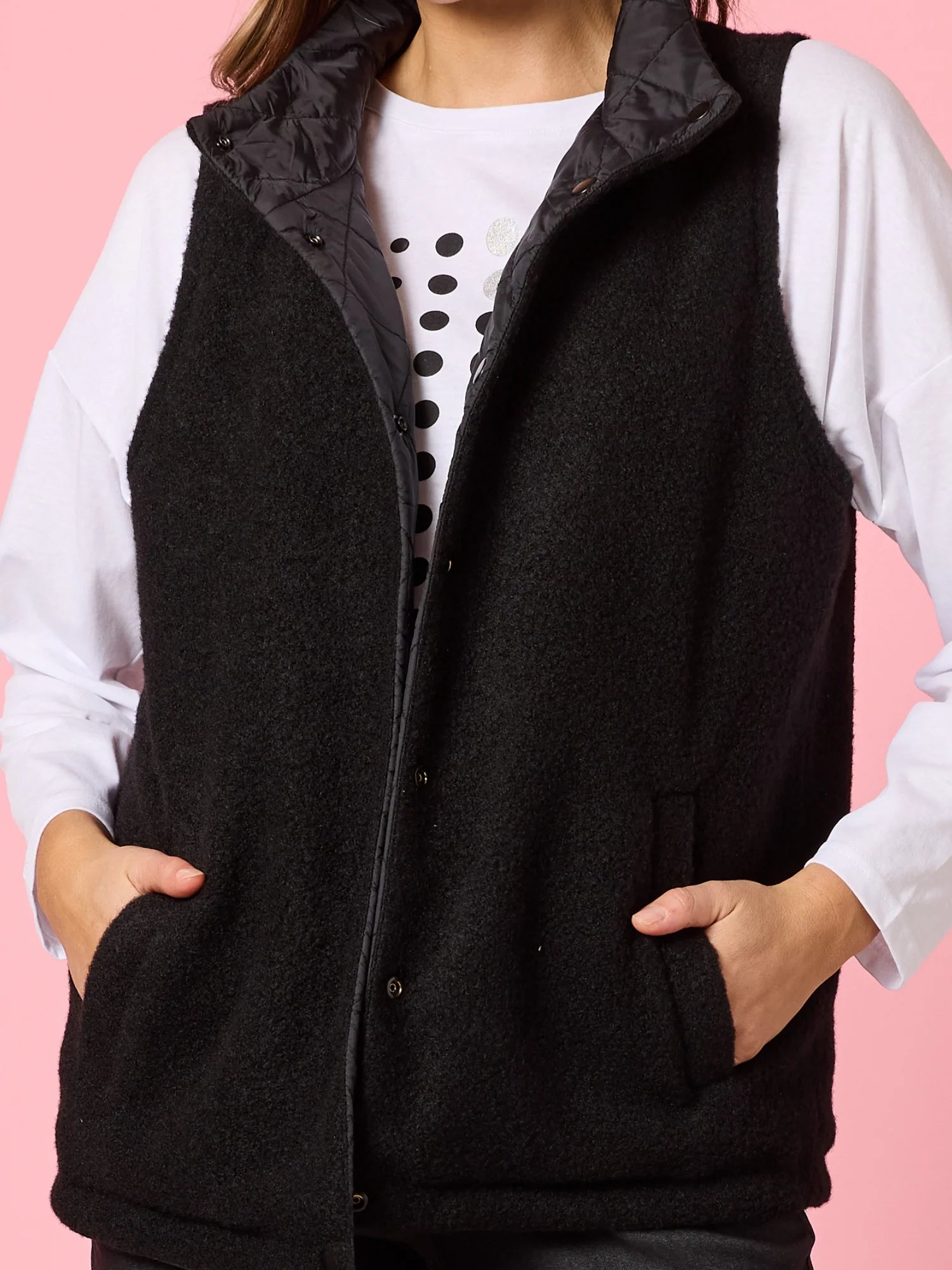 Image of Woman wearing a black vest over a white shirt with polka dots on a pink background