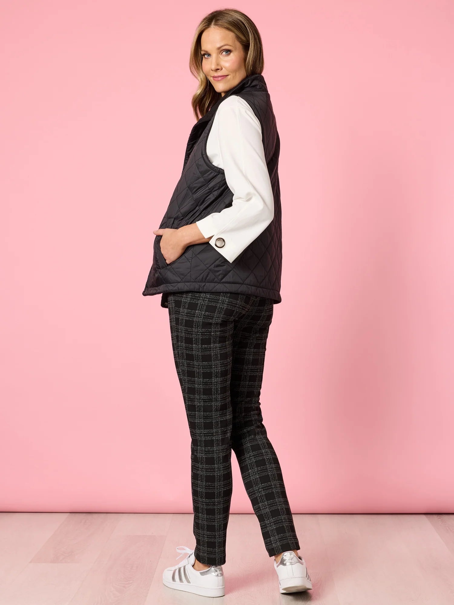Woman wearing a Clarity black vest, white shirt, and plaid pants against a pink background
