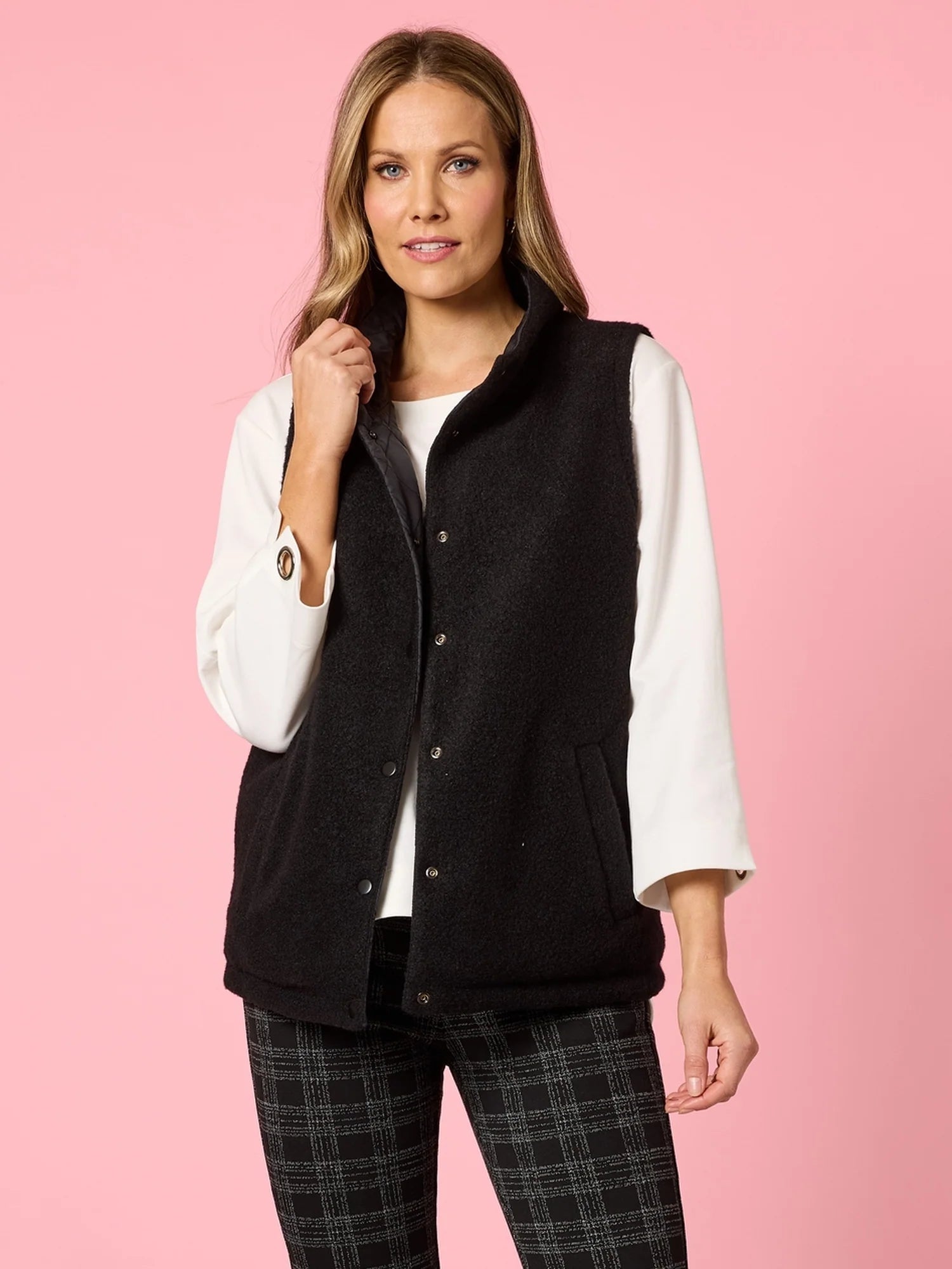 Woman wearing a Clarity black vest over a white shirt with plaid pants on a pink background