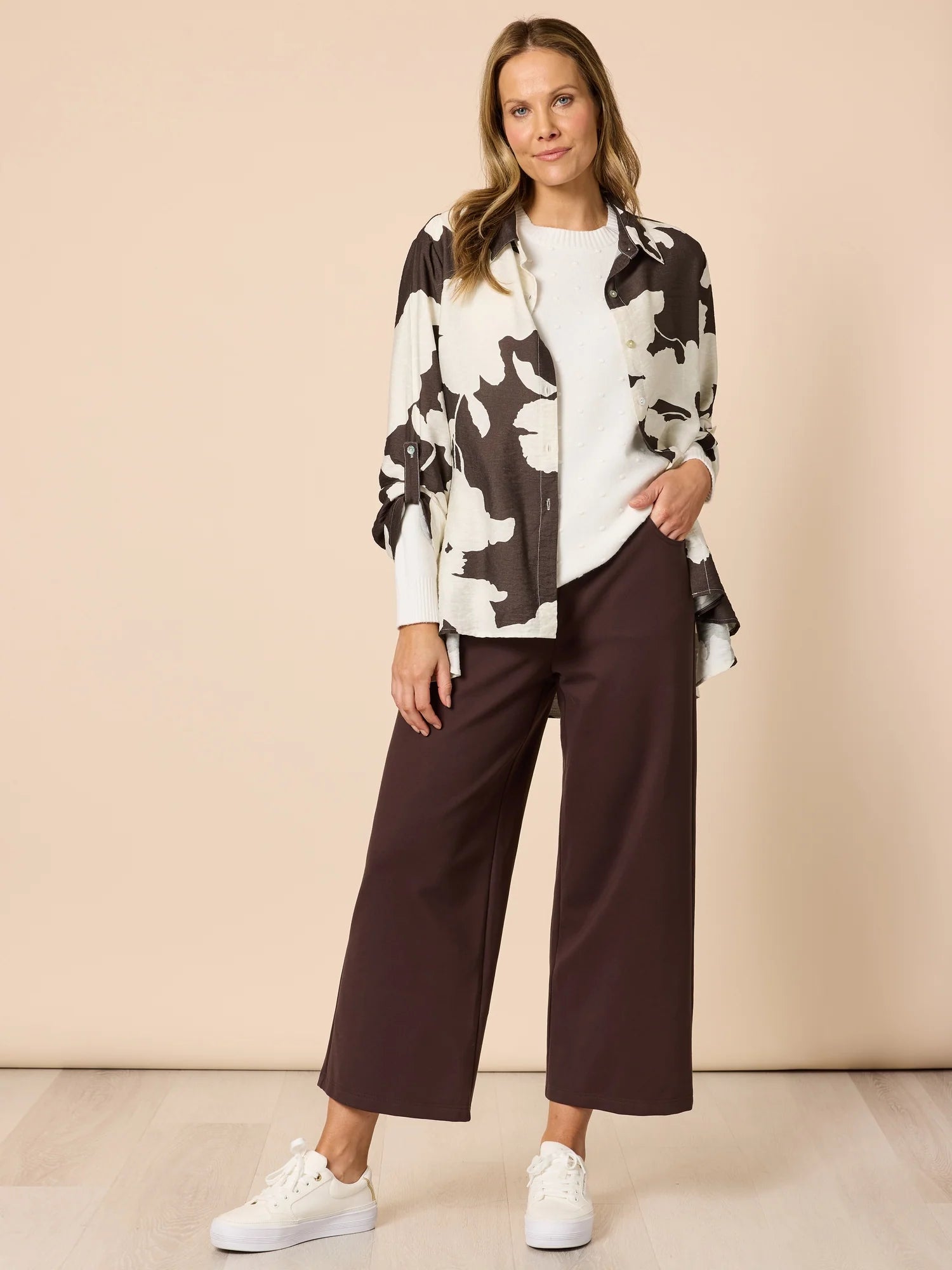 Woman wearing a Clarity black and white floral jacket, white shirt, brown pants, and white sneakers on a beige background