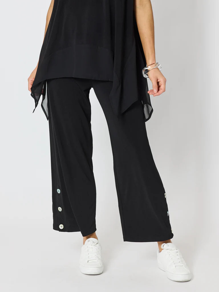 Image of A woman wearing Clarity black wide-leg cropped pants with decorative white buttons down the sides, paired with a black draped top and white sneakers, photographed against a light gray studio background.