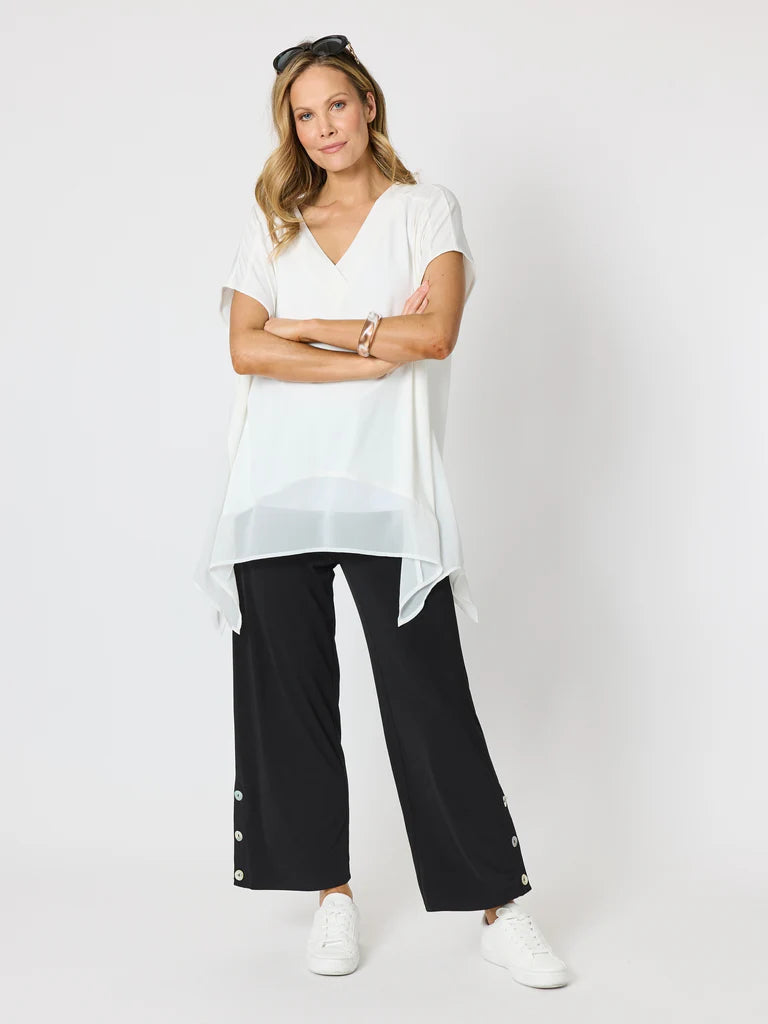 A woman modeling a white short-sleeve V-neck top with a sheer overlay and Clarity black wide-leg pants, standing against a light gray studio background.