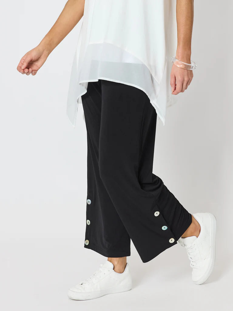 Image of A woman wearing Clairty loose black cropped pants with decorative white buttons at the cuffs, paired with a white sheer overlay top and white sneakers, photographed against a light gray studio background.