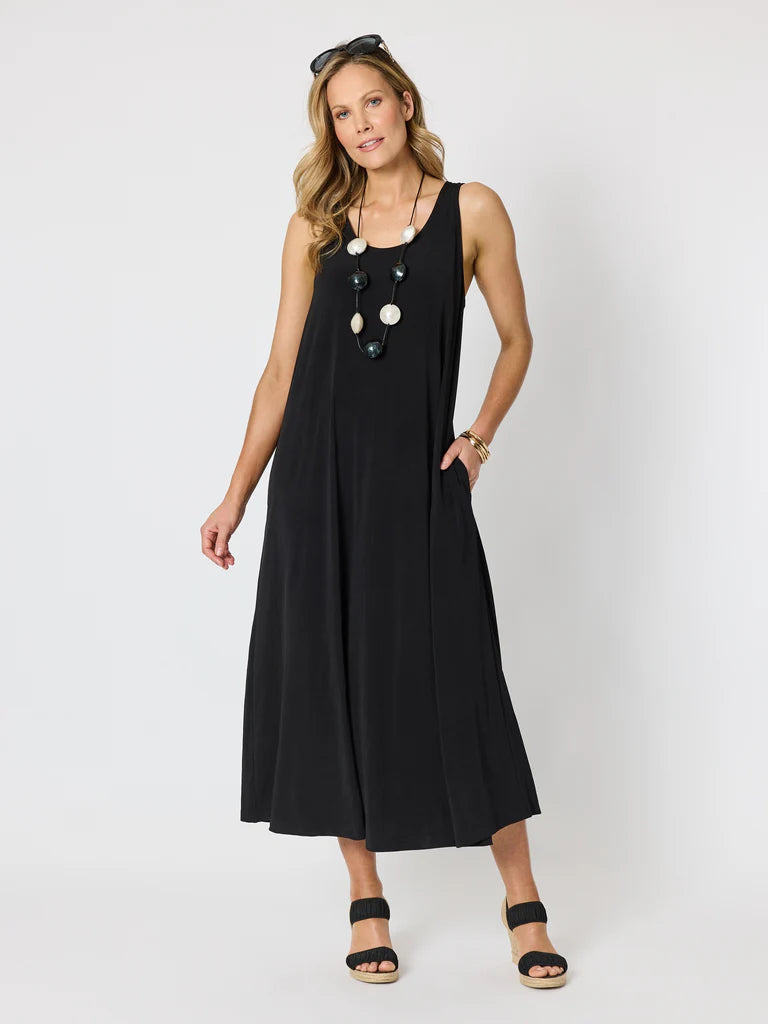 Image of A woman modeling a Clarity sleeveless black midi dress with a decorative multi-colored beaded necklace at the neckline, black sandals, and sunglasses on her head, set against a plain light gray studio background.