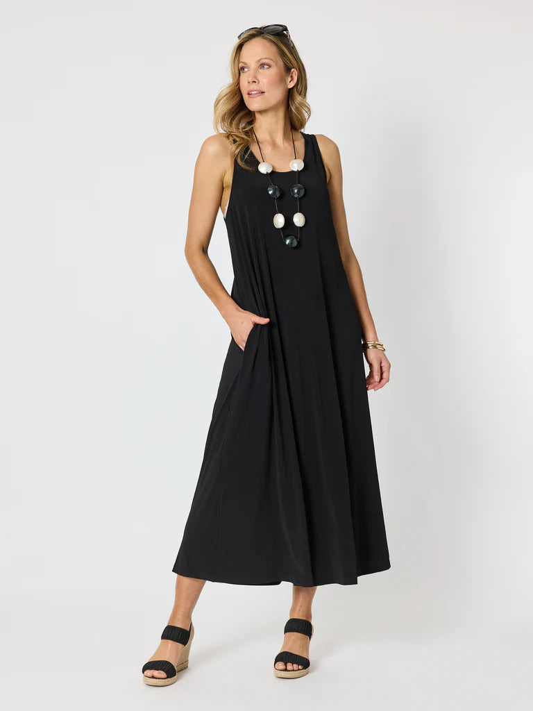 Image of A woman modeling a Clarity sleeveless black midi dress with large decorative white and black buttons down the front, side pockets, and a flowy silhouette, paired with black wedge sandals against a light gray studio background.